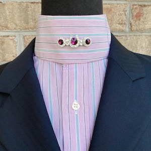 May include: A pink and blue striped dress shirt with a white button and a silver horseshoe pin with purple rhinestones.