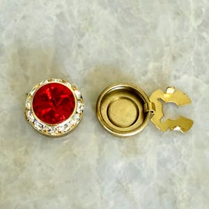 May include: A gold-coloured, round jewellery finding with a large, faceted red stone centre, surrounded by clear crystals. The finding is open, revealing a gold-coloured interior and a hinged clasp.