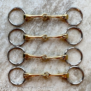 Peut inclure: Quatre porte-clés en métal doré en forme de mors de cheval disposés en colonne verticale. Chaque porte-clés présente une barre centrale avec une boucle à chaque extrémité, reliée à un anneau fendu en métal argenté. Texte : Copyright Show Ring Outfitters.