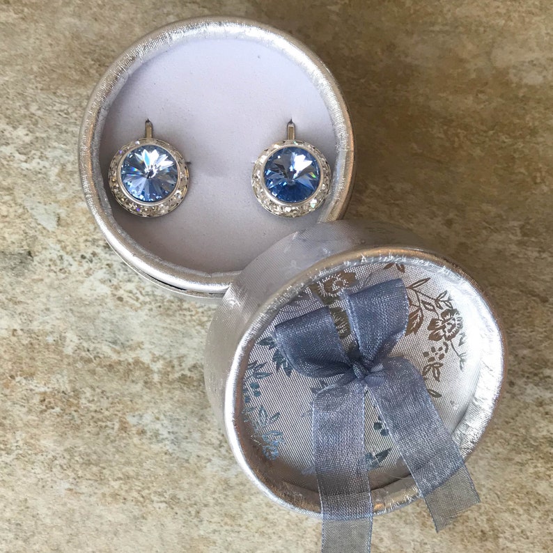 Puede incluir: Un par de pendientes de plata con piedras de cristal azul claro en una caja de regalo plateada con una cinta azul.
