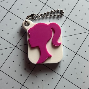 May include: A white rectangular keychain featuring a raised hot pink silhouette of a ponytail hairstyle profile. The keychain has a small circular loop at the top attached to a silver-colored metal ball chain for hanging or attachment purposes.