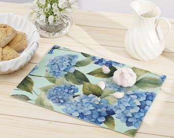 May include: Rectangular glass cutting board featuring a watercolor design of blue hydrangea flowers and green leaves on a light green background. A bulb of garlic and cloves are on the board. The board is on a light wood surface.