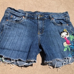 May include: Blue denim shorts with a frayed hem. The shorts feature a Betty Boop graphic on the front, with embroidered flowers and leaves in shades of pink, green, and blue. The shorts have a button and zipper closure.