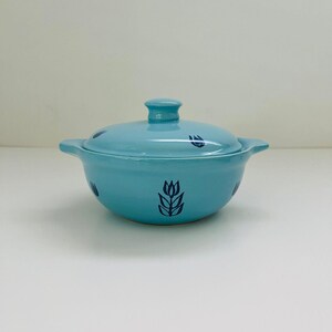 May include: A light blue ceramic casserole dish with a matching lid and two side handles. The exterior of the bowl and the lid are decorated with a repeating dark blue tulip flower pattern. The surface has a smooth, glossy finish throughout.
