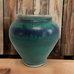 May include: A handcrafted ceramic vase with a wide, rounded body and a slightly flared rim. The vase features a gradient of colors, transitioning from a deep teal at the base to a vibrant green and a band of dark blue near the top. The rim is a lighter, muted green.