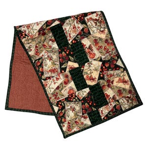 May include: A rectangular table runner featuring a patchwork design of cream-colored squares with illustrations of fairies and red berries, alternating with solid dark green squares. The reverse side is a solid, textured terracotta-colored fabric.