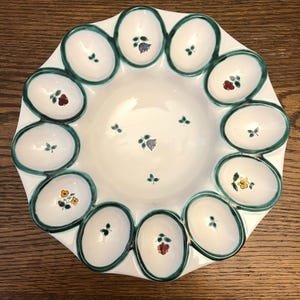 May include: A white ceramic egg serving platter with a scalloped edge and a green border. Each egg-shaped section is decorated with a small floral design in various colors. The center of the platter also features floral accents.