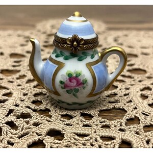May include: A miniature porcelain teapot trinket box featuring a white base with hand-painted pink rose motifs, green leaf garlands, and light blue panels. The lid is hinged with a brass flower-shaped clasp and gold-painted trim on the spout and handle.