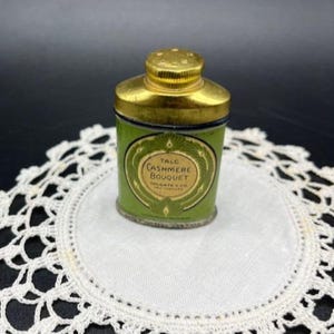 May include: Vintage green talc powder tin with a gold lid. The tin features the text "Talc Cashmere Bouquet Colgate & Co" in a decorative oval. The tin is set on a white lace doily.