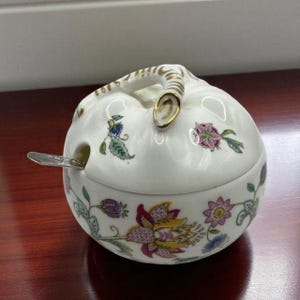 May include: A white ceramic sugar bowl with a lid and handle, adorned with floral patterns in pink, purple, and blue. A small silver spoon rests in the bowl. The handle and rim have gold accents.