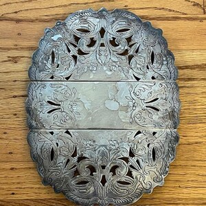 May include: An antique silver-colored trivet with an ornate, openwork design. The trivet is oval-shaped and segmented into three hinged sections. The decorative pattern includes floral and scroll motifs, adding a vintage aesthetic to the kitchenware.