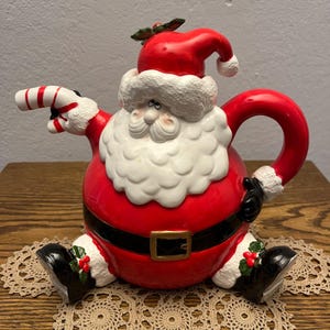 May include: A ceramic Santa Claus teapot featuring a round red body, a black belt with a gold buckle, and a white beard. The spout is shaped like a red and white candy cane, and the handle is red. Santa wears a red hat with white trim.