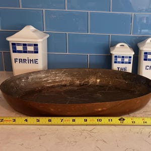 May include: A well-worn, copper-colored frying pan with a long, dark handle. The pan is approximately 14 inches in diameter. Three white ceramic canisters with blue checkered patterns and labels reading "FARINE", "THE", and "CAFÉ" are in the background.
