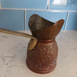 May include: A copper coffee pot with a long brass handle and a wide pouring spout. The body of the pot features an embossed floral pattern throughout. The interior of the pot shows a metallic finish, and the exterior has a warm, aged copper tone.