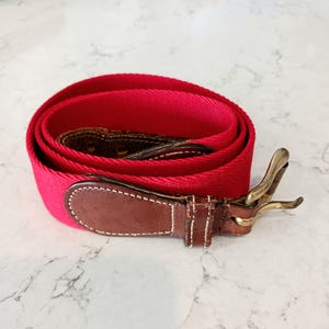 May include: A rolled-up red canvas belt with a brown leather accent and a brass buckle. The leather accent is stitched with white thread. The belt is a classic accessory.