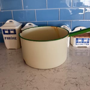 May include: A cream-colored enamel saucepan with a green handle and rim. The pot is set on a countertop with blue tile in the background. Two white containers with blue checkered patterns and text are also visible.