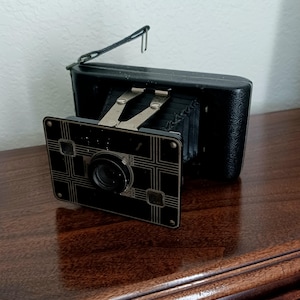 May include: A vintage black camera with a decorative front panel featuring a geometric design. The camera has a collapsible bellows and a metal viewfinder. The camera is resting on a dark wooden surface.