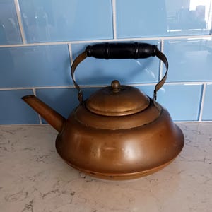 May include: A copper-colored tea kettle with a black wooden handle and a curved spout. The kettle has a rounded body and a small lid with a knob. The handle is attached to the kettle with brass-colored metal. The background is a light blue tile.