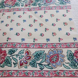 May include: Cream-colored fabric with a floral pattern. The design features large flowers in pink, blue, and red, with green leaves, set against a cream background. The fabric has a striped border with smaller floral accents.