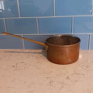 May include: A round copper saucepan with a long, straight brass handle attached by a metal rivet. The interior of the pan shows a metallic finish, while the exterior displays a warm, weathered copper patina with visible signs of use.