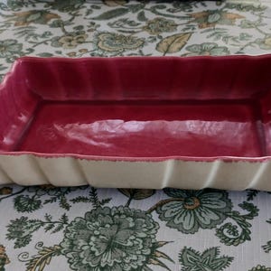May include: A rectangular ceramic serving dish with a deep red interior and a cream-colored, ridged exterior. The dish has a slightly scalloped edge and is likely used for serving food.