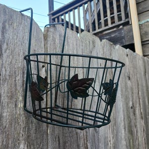 May include: A dark green metal hanging basket with leaf accents, attached to a wooden fence. The basket is designed to hold plants and has a decorative leaf pattern. The fence is made of weathered wood.