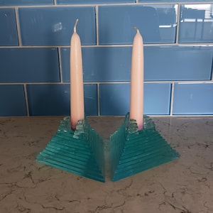 May include: Two teal glass candle holders with a stepped, pyramid-like design. Each holder features a central opening holding a pale pink taper candle. The glass has a translucent, layered appearance with a jagged, textured top edge.