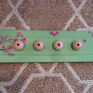 May include: A light green wooden wall-mounted coat rack with four pink and white decorative knobs. The rack is adorned with pink cherry blossom branches. A charming and functional piece for any entryway or bedroom.