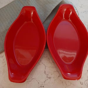 May include: Two bright red, oval-shaped serving dishes. Each dish has a slightly raised rim and a small handle-like extension at one end. The dishes are made of a glossy material and are set against a light gray background.