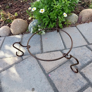 May include: A decorative, circular, brown metal plant stand with three curved legs and a unique, artistic design. The stand is designed to hold a potted plant. The stand is placed on a gray brick patio, with a background of flowers and rocks.