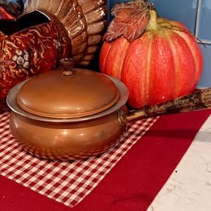 May include: A small copper saucepan with a lid, featuring a wooden handle. The pot sits on a red and white checkered cloth, with a decorative pumpkin and turkey figurine in the background. The handle is approximately 6 inches long.