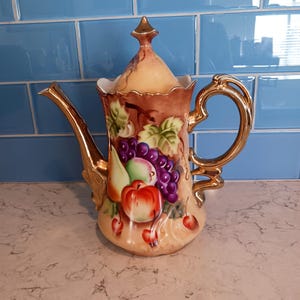 May include: A vintage ceramic teapot with a gold handle, spout, and accents. The teapot is decorated with a colorful fruit design, including grapes, apples, and pears. The background is a light beige with a blue tile backdrop.