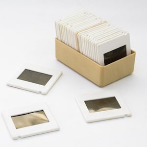 Puede incluir: Una pila de monturas de diapositivas de plástico blanco en una caja beige, con tres monturas de diapositivas adicionales sobre una superficie blanca.