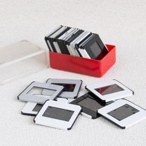 Puede incluir: Una caja de plástico roja llena de una pila de negativos de película de diapositivas en blanco y negro. Una caja de plástico transparente y una pila de negativos de película de diapositivas están sobre una superficie blanca.