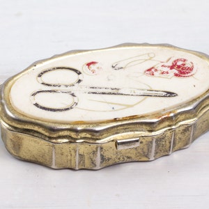 Può includere: Una scatola vintage ovale in metallo con interno bianco e bordo dorato. Il coperchio presenta un disegno di forbici, filo e un fiore in rosso e bianco.