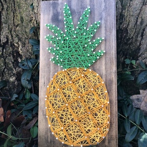 Op de afbeelding: Een string art ananas met een groene bovenkant en een geel lichaam op een houten achtergrond.