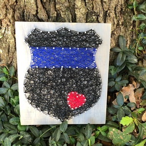 May include: A string art piece depicting a police badge with a blue stripe and a red heart in the center. The badge is made of black string wrapped around nails hammered into a wooden board.