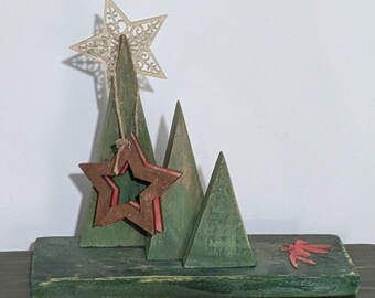 May include: A wooden Christmas decoration featuring three green triangular trees on a rectangular base. A decorative star tops the tallest tree, and a red and brown star hangs from the middle tree. A red leaf is on the base.