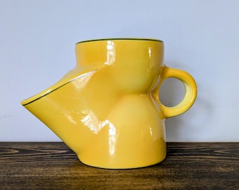 May include: A yellow ceramic pitcher with a unique, curved design. The pitcher has a spout, a rounded handle, and a black trim along the rim and spout. The glossy finish reflects light, highlighting the bright yellow color.