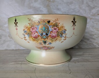 May include: Cream-colored ceramic bowl with a pedestal base. The bowl features a floral design with pink roses, blue accents, and gold trim. The rim has a green gradient, and the bowl is decorated with fleur-de-lis motifs.