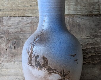 May include: A ceramic vase with a light blue and tan gradient. The vase features a hand-painted scene of figures under a tree with birds in flight. The vase has a flared top and a rounded body, perfect for displaying flowers.