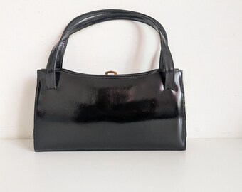 May include: A glossy black handbag with two handles and a rectangular shape. The bag has a gold-colored clasp and a shiny, reflective surface. The handles are thin and attached to the top of the bag.