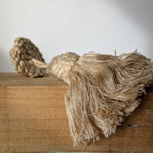 May include: A decorative tassel in a light beige hue, featuring a woven top and a flowing, fringed bottom. It is positioned on a wooden surface. This tassel is suitable for home decor or craft applications.