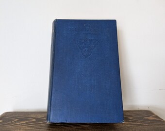 May include: A vintage, navy blue book with embossed text and a crest design on the cover. The book is standing upright on a wooden surface against a white background. The book's title is visible.