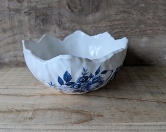 May include: A white ceramic bowl with a scalloped rim and blue floral pattern. The bowl is round and sits on a wooden surface. The blue floral design is on the outside of the bowl.