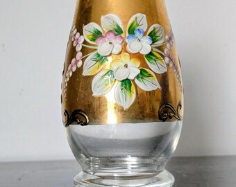 May include: A clear glass vase with a gold band featuring hand-painted floral designs in white, pink, and blue. The vase has a flared top and a round base, with black decorative accents.