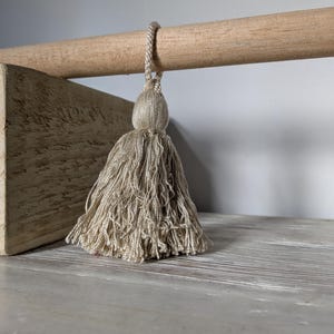 May include: A beige tassel suspended from a wooden dowel. The tassel is made of multiple strands of thread, and the dowel is supported by a light-coloured wooden block. The background is a neutral grey.