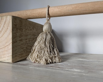 May include: A beige tassel hangs from a wooden dowel. The tassel is made of multiple strands of thread, and the dowel is supported by a light-colored wooden block. The background is a neutral gray.