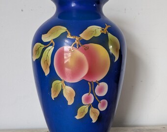 May include: A cobalt blue vase with a glossy finish. The vase is decorated with a painted design of peaches and leaves in shades of pink, yellow, and green. The fruit and foliage are arranged on a gold-colored branch.