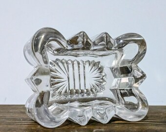 May include: Clear, rectangular glass ashtray with a decorative, scalloped edge. The center features a sunburst design. The ashtray sits on a wooden surface against a white background. The glass has a reflective quality.